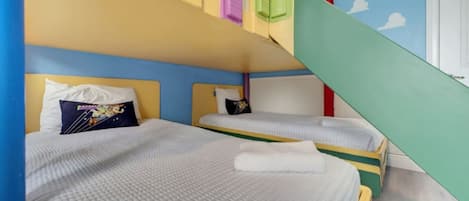 A room with two beds, each with a pillow and a blanket. There is a staircase with colorful steps leading to a door.