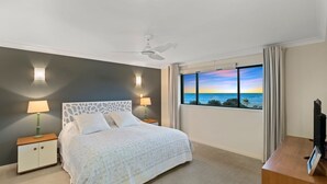 A bedroom with a bed, two bedside tables, a ceiling fan, a TV, and a window with a view of the ocean.