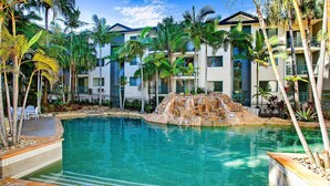 A swimming pool surrounded by palm trees and a multi-story building with balconies.