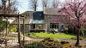 A stone house with a conservatory, a wooden pergola, and a garden with a lawn and flowering tree. - The Old Coach House (Alnwick)