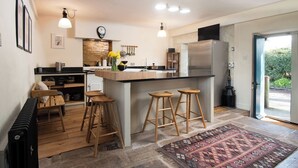 A modern kitchen with a central island, bar stools, a refrigerator, and a dining area with a bench. - The Old Coach House (Alnwick)