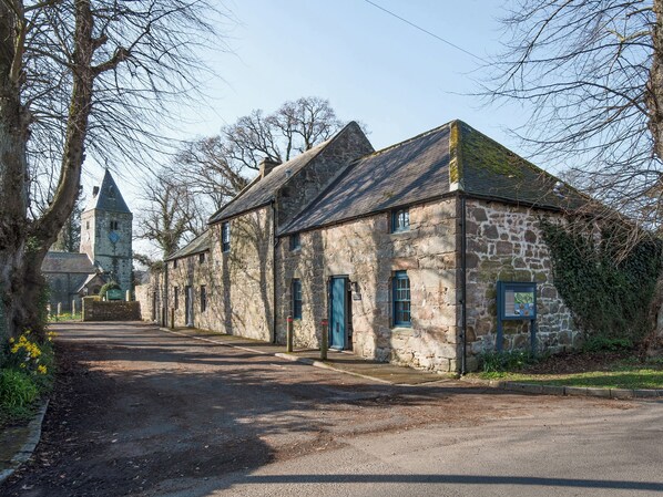 A stone building with a traditional architectural style, a church tower in the background, and a paved road. - The Old Coach House (Alnwick)