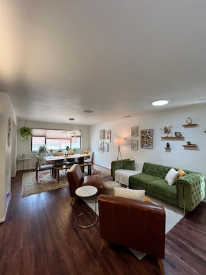 A living room with a green sofa, a brown leather chair, a dining area with a table and chairs, and a kitchen in the background.