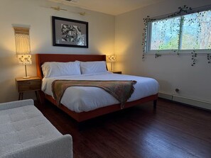 A bedroom with a wooden bed, two bedside lamps, a framed picture on the wall, and a window with greenery.