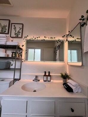 Un baño con tocador blanco, un espejo grande y repisas con toallas y artículos de tocador.