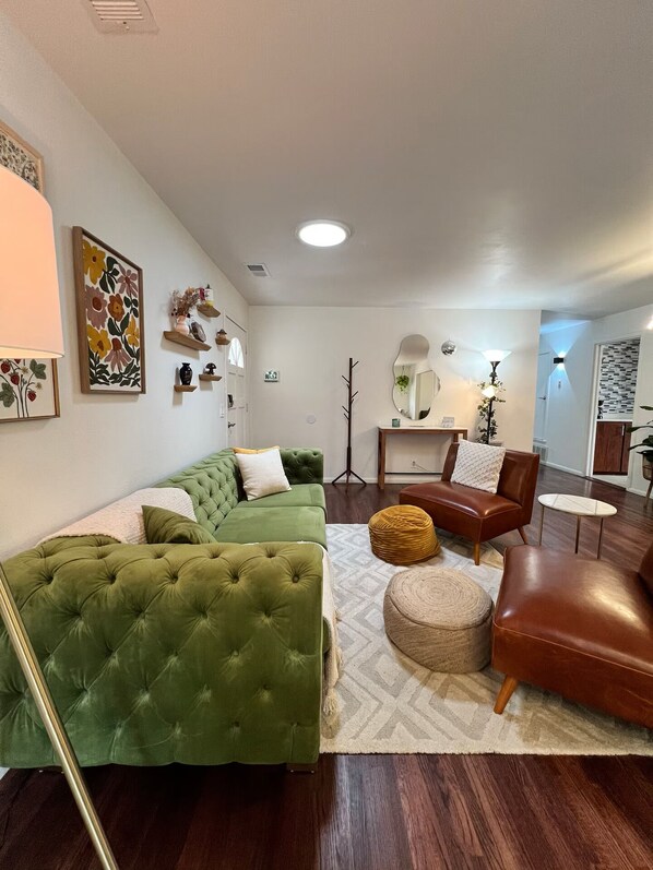 Una sala de estar con un sofá verde, un sillón de cuero marrón, un otoman redondo y una mesita de noche con una lámpara.