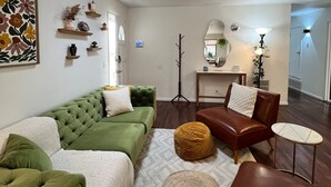 Un salon avec un canapé vert, un fauteuil en cuir marron, un petit pouf rond et une table d’appoint.