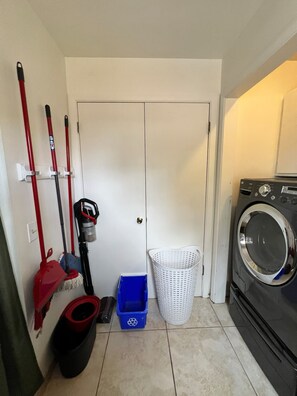 A laundry room with a washing machine, a mop, a broom, a vacuum cleaner, and a laundry basket.