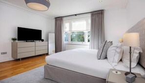 A modern bedroom with a large bed, a television, a dresser, and a window with curtains. - Stylish Three-Bedroom Apartment in the Heart of Belsize Park (London)