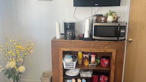 A room with a television mounted on the wall, a microwave on a wooden cabinet, and a coffee maker. There is a small shelf with various items like jars and containers.
