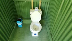 A small, enclosed bathroom with a toilet, a blue trash bin, and a roll of toilet paper on the wall. - Zompopas Trail (Puerto Jiménez)