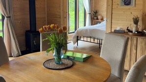 A wooden cabin interior with a bed, a table with a vase of tulips, and a view of greenery outside. - Primrose (Chard)