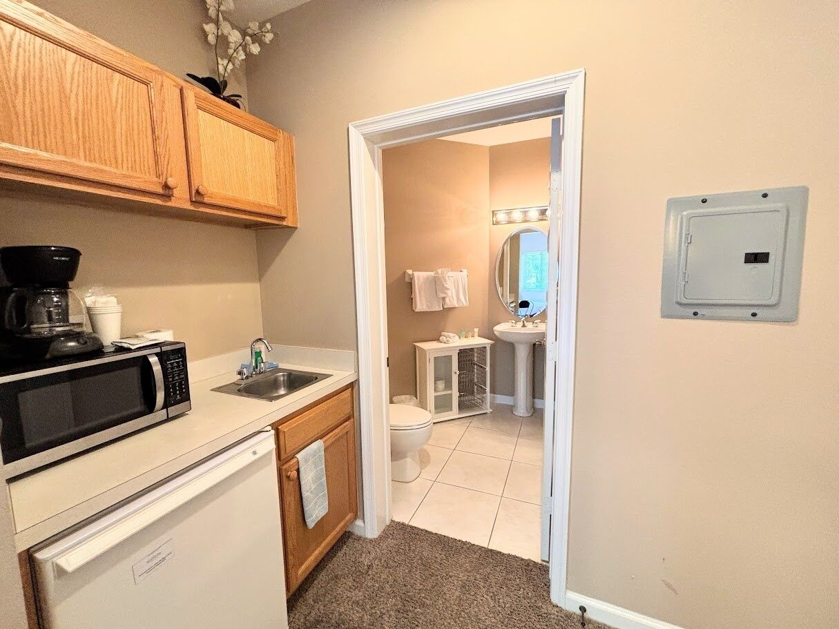 A compact kitchen with a microwave, sink, and cabinets, leading to a bathroom with a toilet and sink.