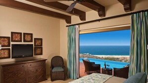 Ein Schlafzimmer mit großem Fenster, das Blick auf Strand und Meer bietet, ein Bett, ein Fernseher und eine Kommode.