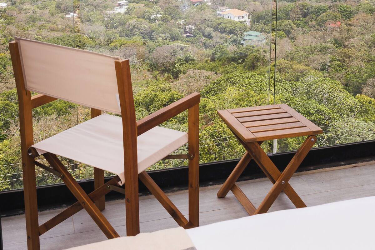A balcony with a wooden chair and a small table, overlooking a coastal landscape with greenery and a view of the sea.
