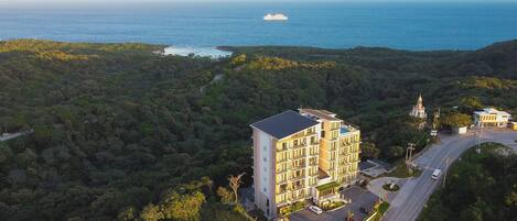 A multi-story hotel with a rooftop terrace, surrounded by dense forest and a view of the ocean.