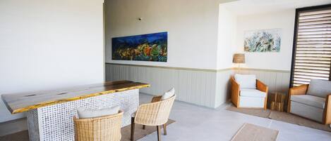 A modern living room with a wooden bar, wicker chairs, and a large abstract painting on the wall.