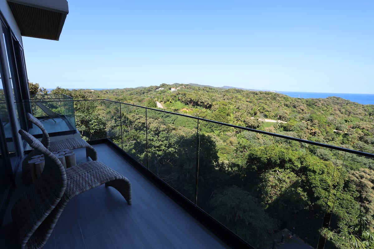 A balcony with wicker furniture, a glass railing, and a view of a lush, green landscape with hills and a distant coastline.