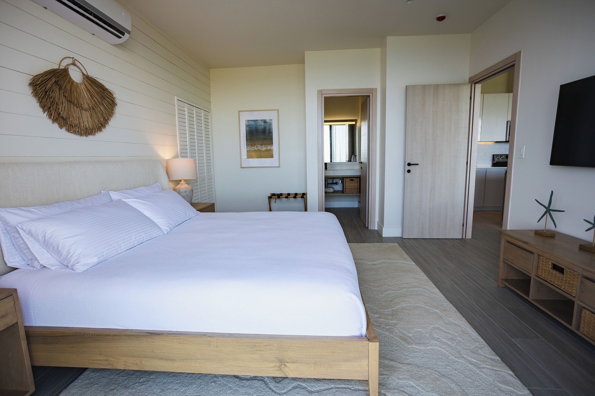 A bedroom with a large bed, a bedside table, a wall-mounted air conditioner, a framed picture, and a wooden nightstand.