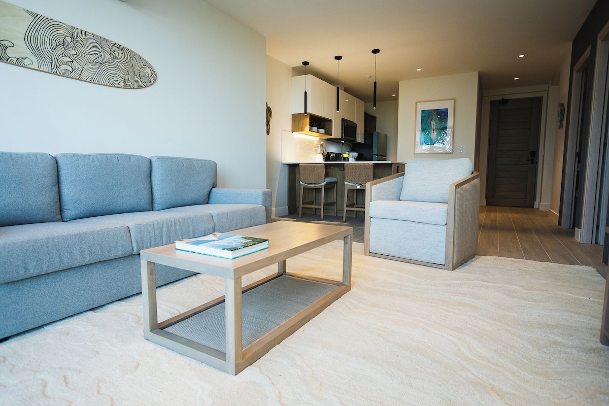 A modern living room with a sofa, a coffee table, and a chair. There is a kitchen area with a TV and bar stools.