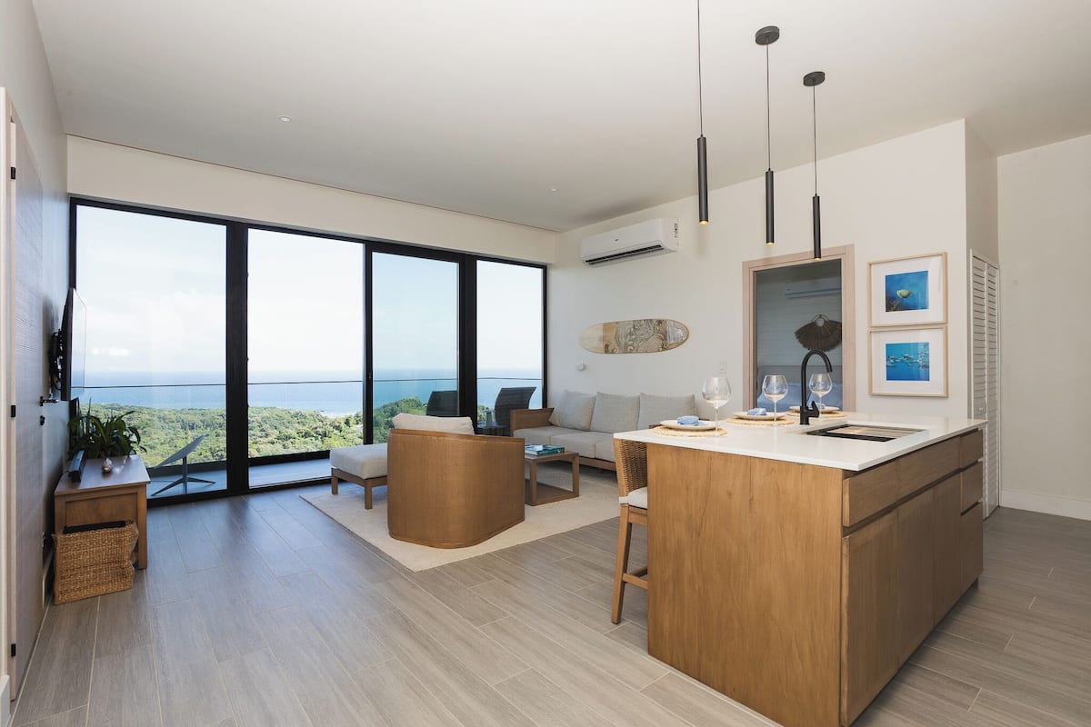A modern living room with a large window, a sofa, a coffee table, and a wooden island with a sink.