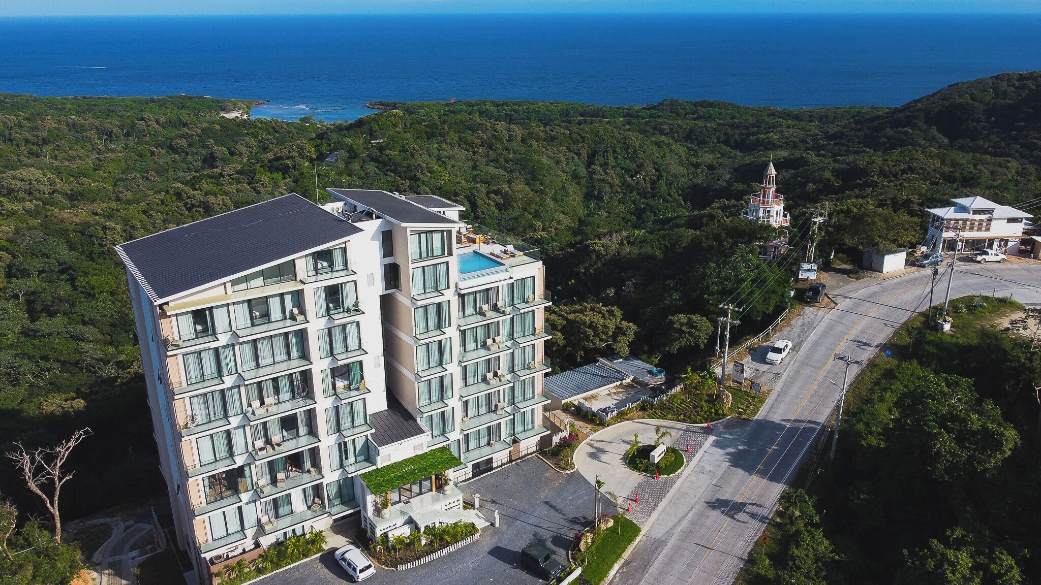 A multi-story hotel with a rooftop pool, surrounded by lush greenery and a view of the ocean.