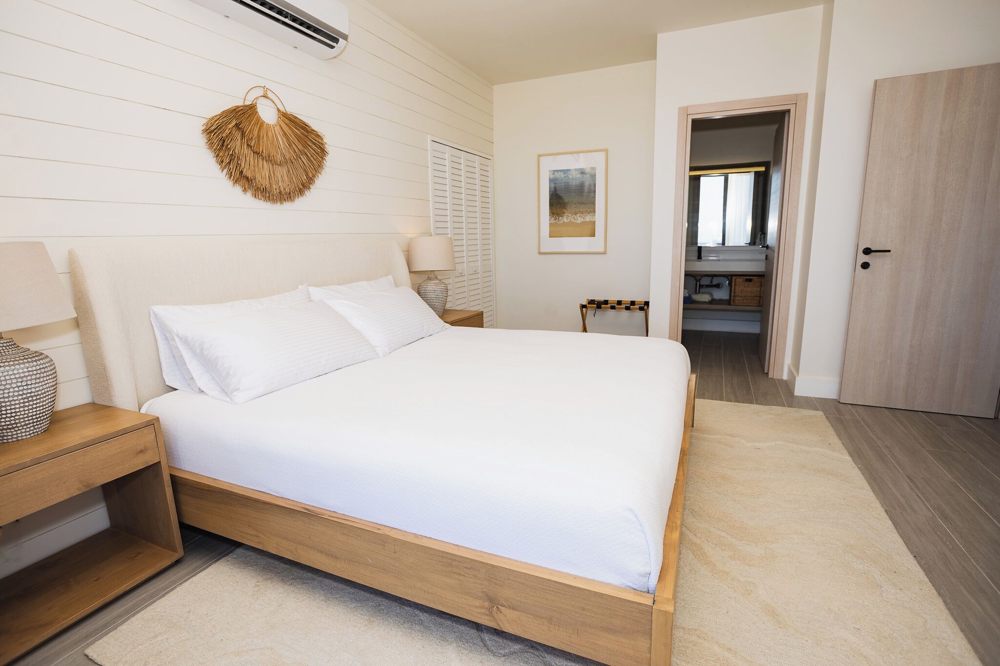 A bedroom with a wooden bed, a bedside table, a hanging wall decoration, a framed picture, and a door leading to another room.