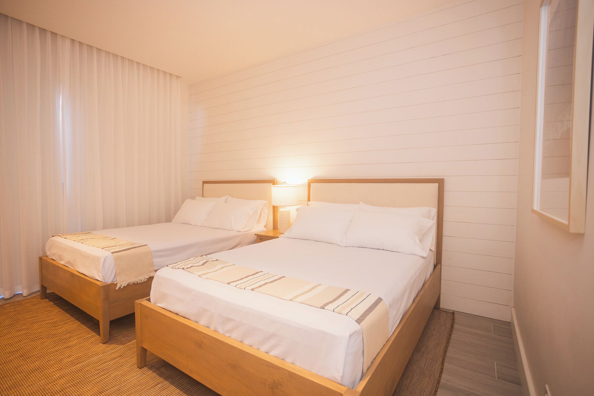 A hotel room with two single beds, a large window with curtains, a wooden floor, and a wall-mounted light fixture.