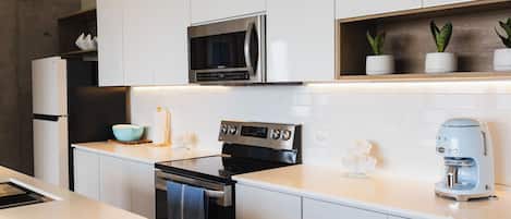 A modern kitchen with white cabinets, a stainless steel microwave, a black oven, a coffee maker, and a small plant on the shelf.