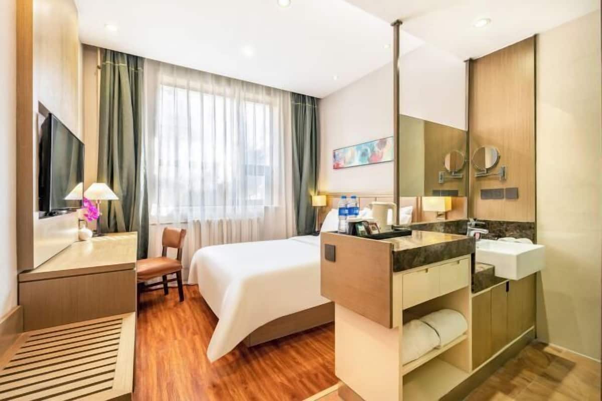 A hotel room with a bed, a desk with a lamp, a chair, a large window with curtains, a bathroom with a sink and mirror, and a flat-screen TV.