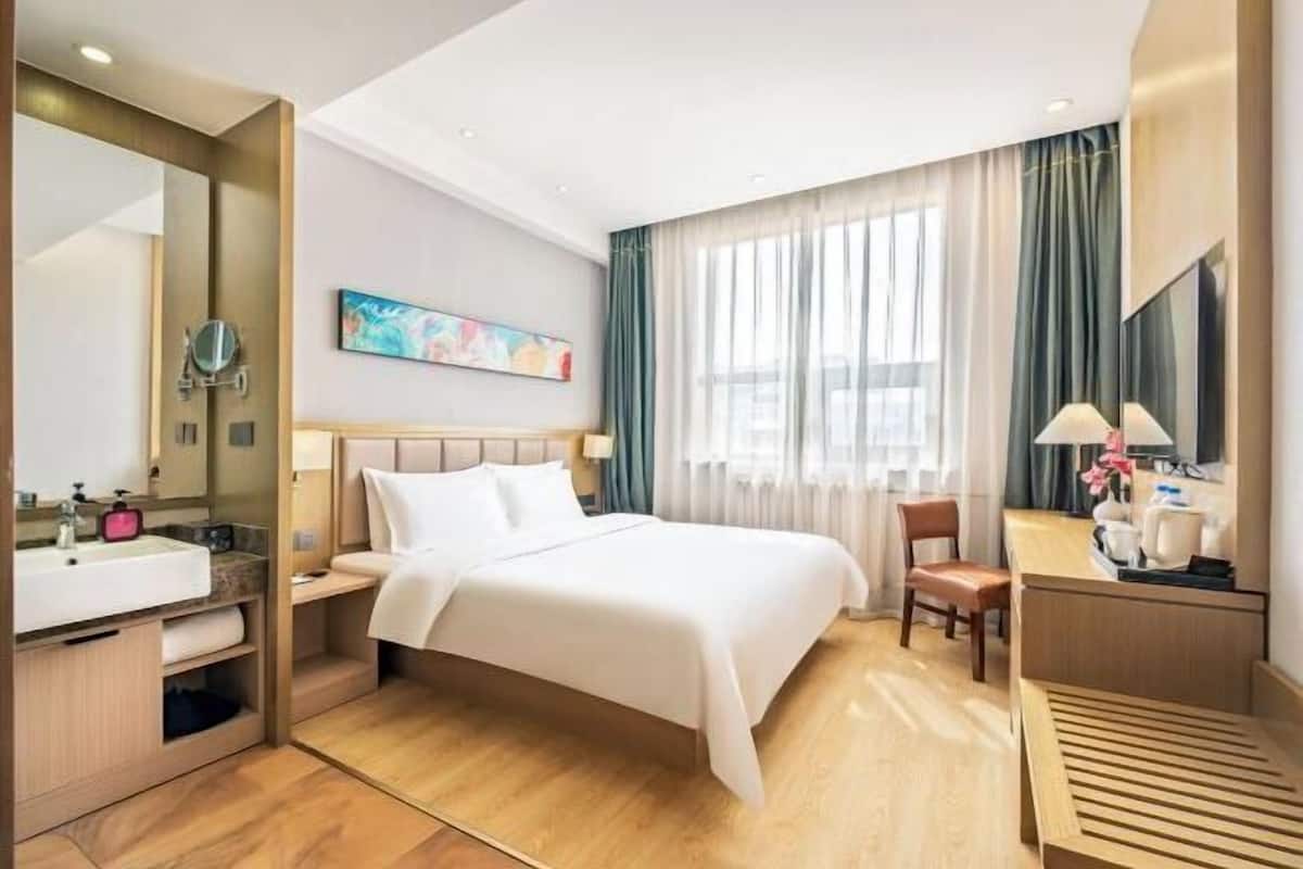 A hotel room with a bed, a desk, a chair, a large mirror, a sink, and a window with curtains.