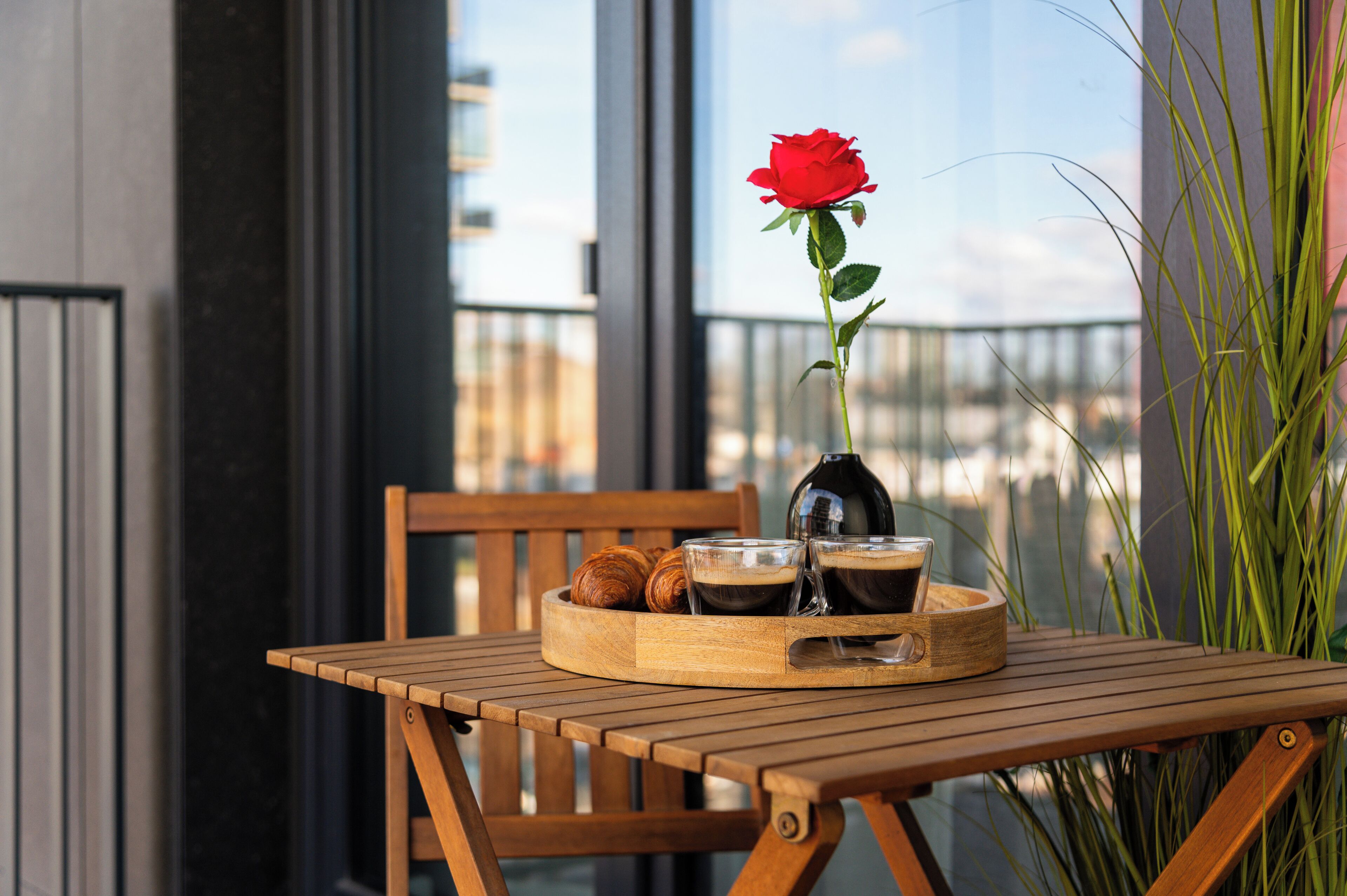 Une table en bois sur laquelle se trouve un plateau contenant des croissants, des tasses de café et une rose rouge.