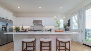 Cocina moderna con armarios blancos, un refrigerador de acero inoxidable, una isla grande con taburetes y vistas al exterior a través de ventanas amplias.