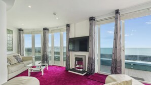 A spacious living room with a large TV, a fireplace, and a balcony with a view of the sea. - Blue Horizon (Lymington)
