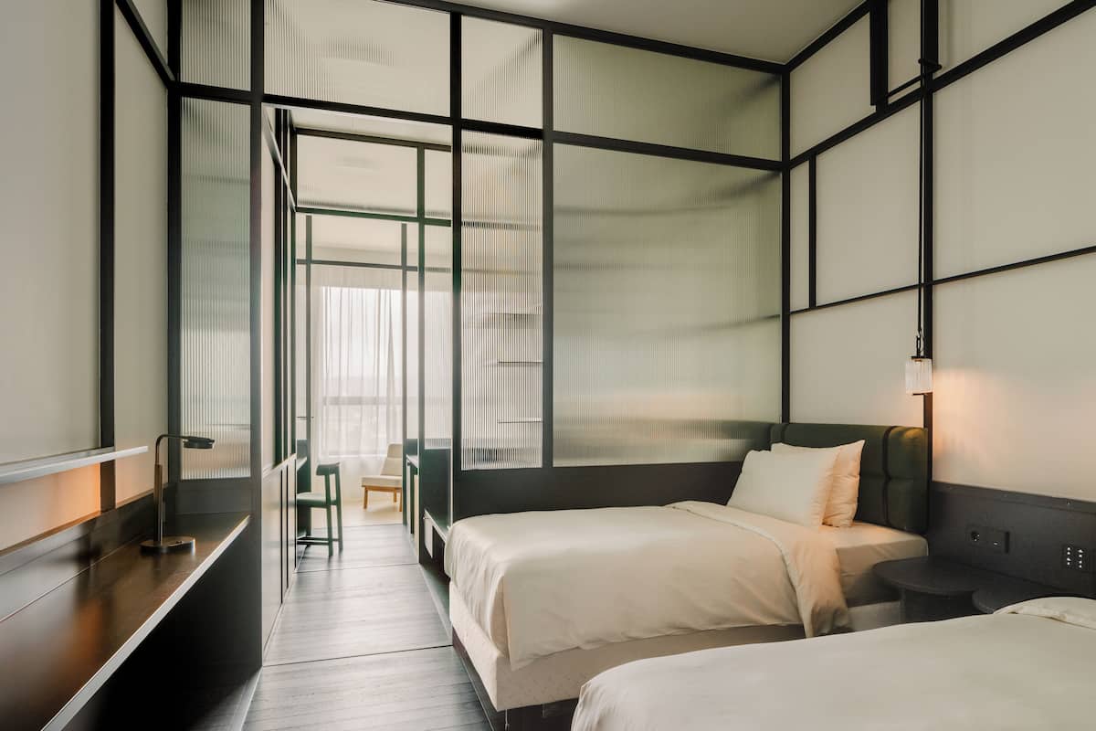 A hotel room with two beds, a desk, a chair, and a view through a large window.