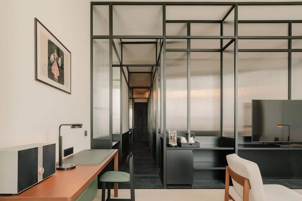 A modern hotel room with a wooden desk, a green chair, a television, and a large window with a view.