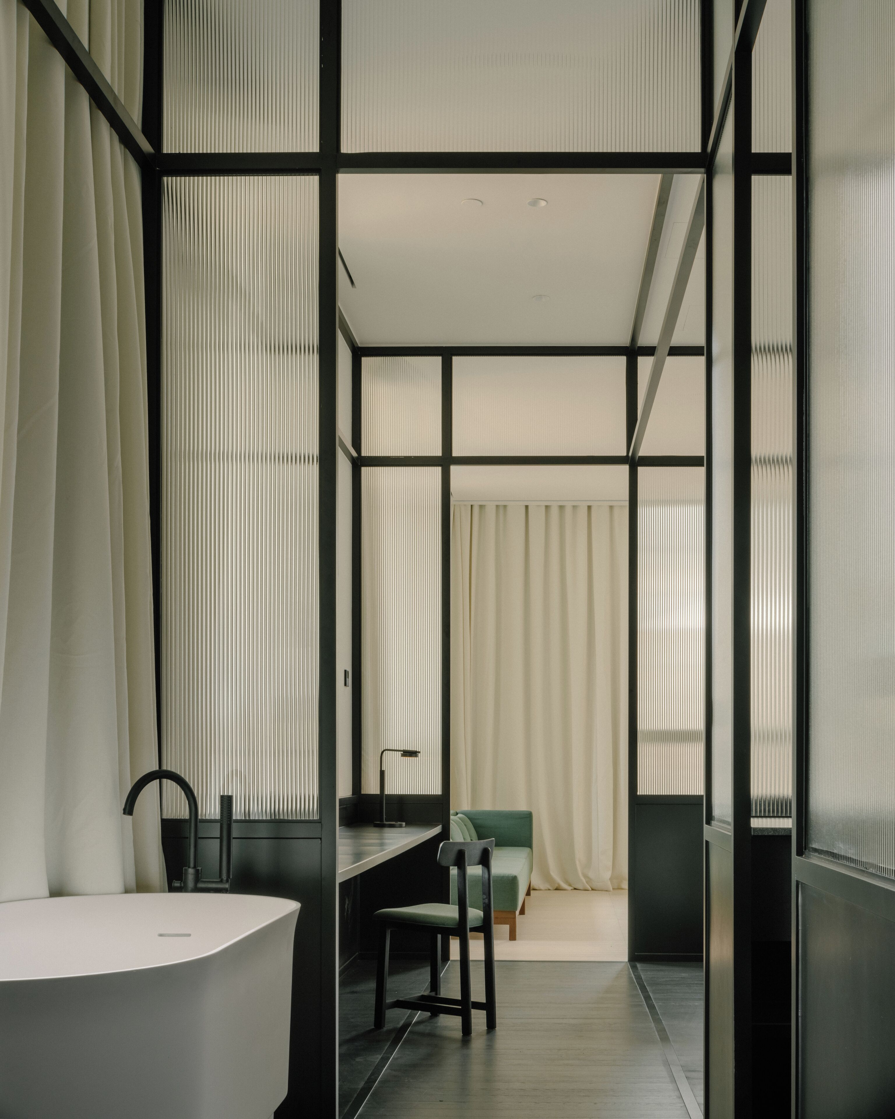 A modern, minimalist bathroom with a large white bathtub, a black sink, and a green chair.