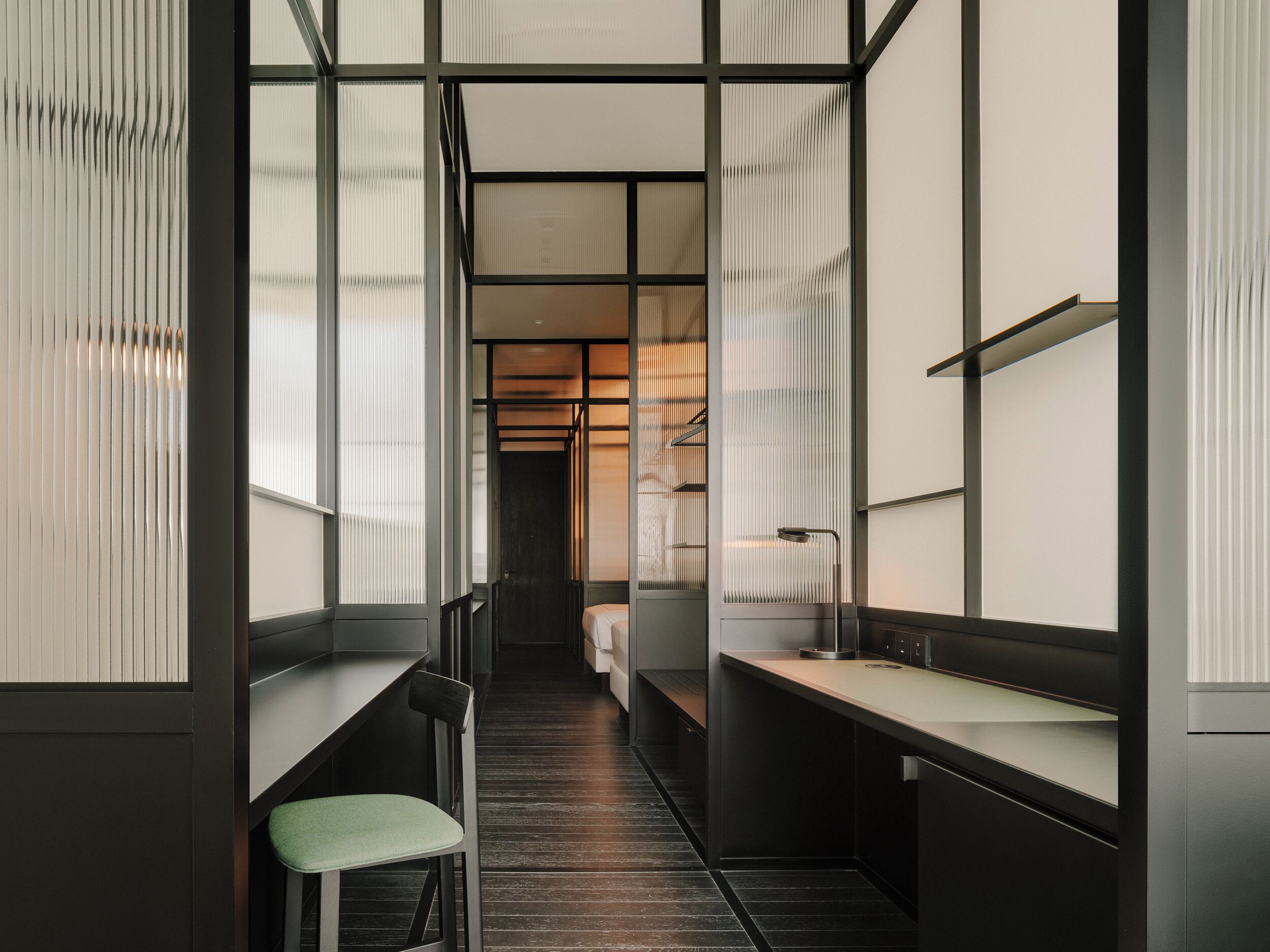 A narrow hallway with a bench and a chair, leading to a bed in a room with a window and a shelf.