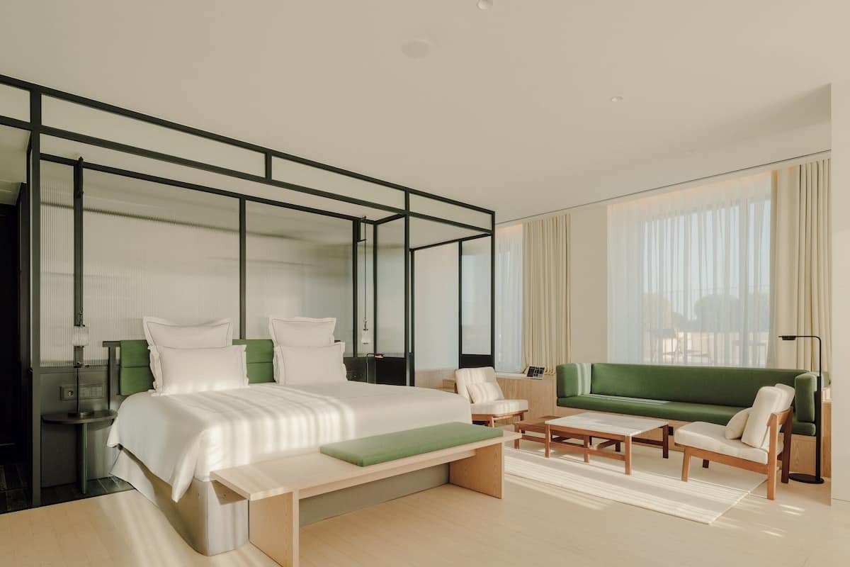 A modern hotel room with a large bed, a green sofa, a wooden coffee table, and a bench.