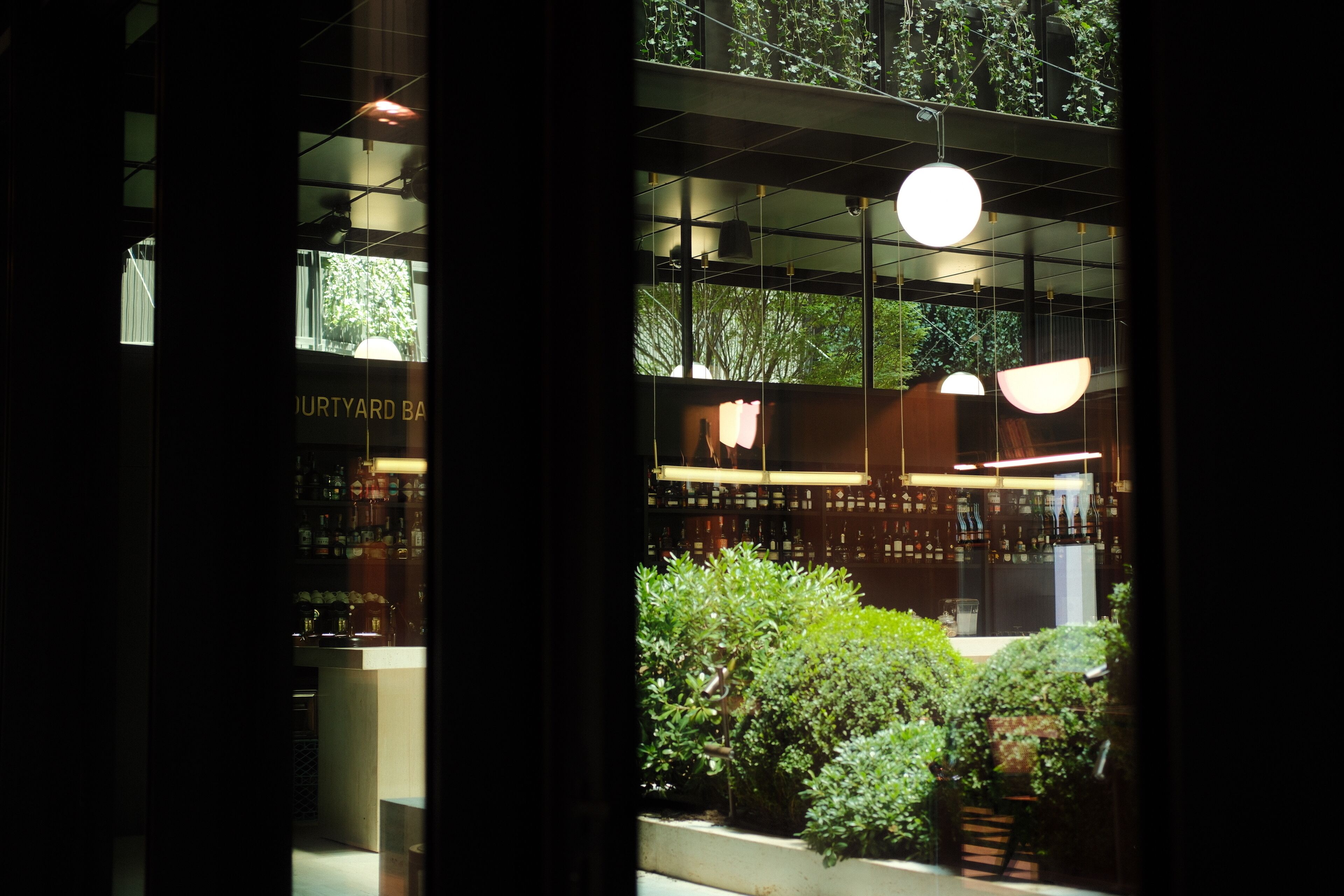 Een uitzicht op een bar met flessen op de planken, hangende lampen en planten zichtbaar door het raam.