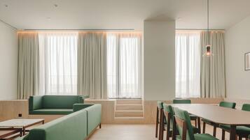 A modern dining area with green upholstered seating, wooden tables, and large windows with sheer curtains.