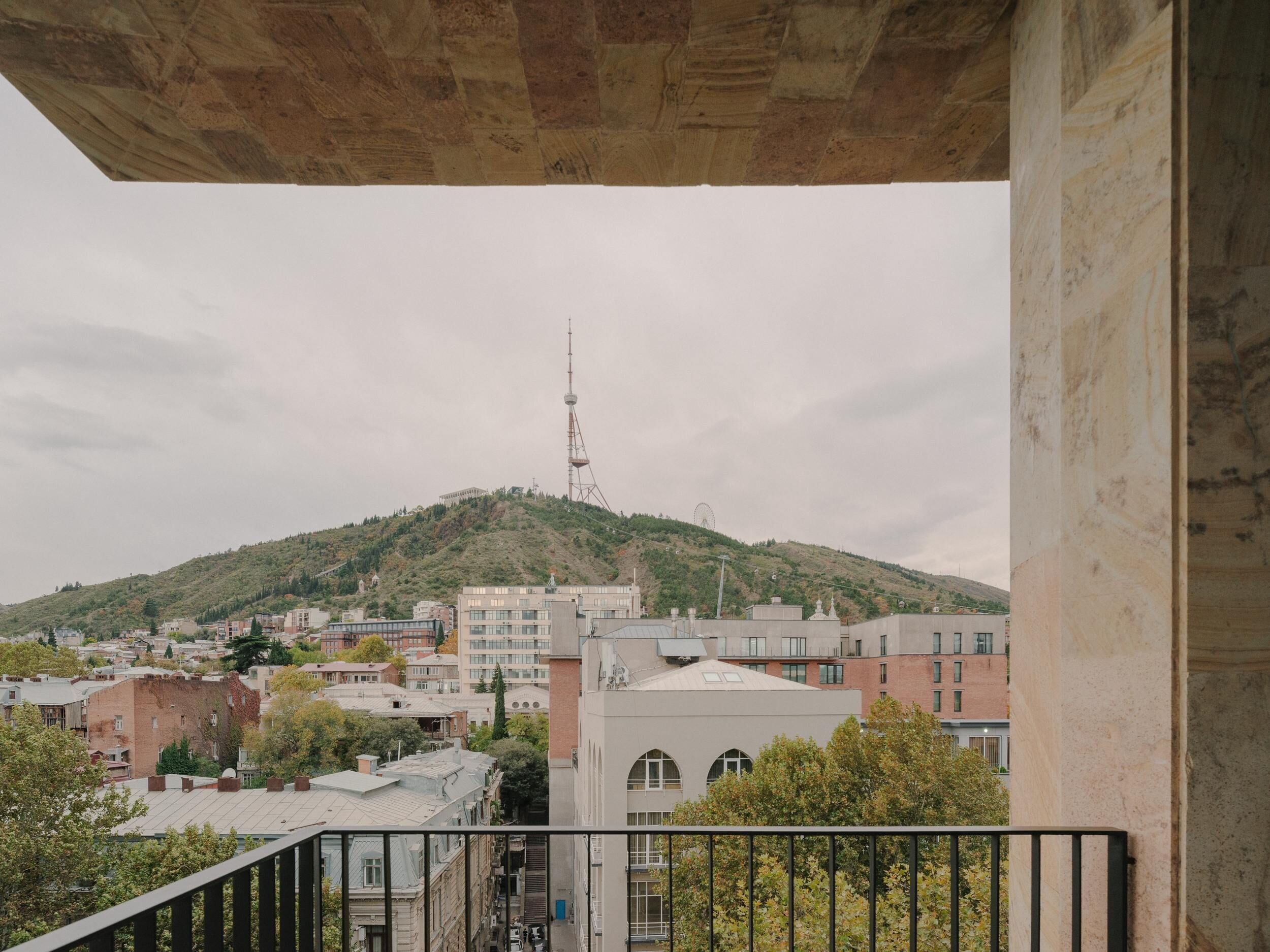 Vista desde un balcón con vistas a un paisaje urbano que incluye edificios, una colina con una torre y una noria a lo lejos.