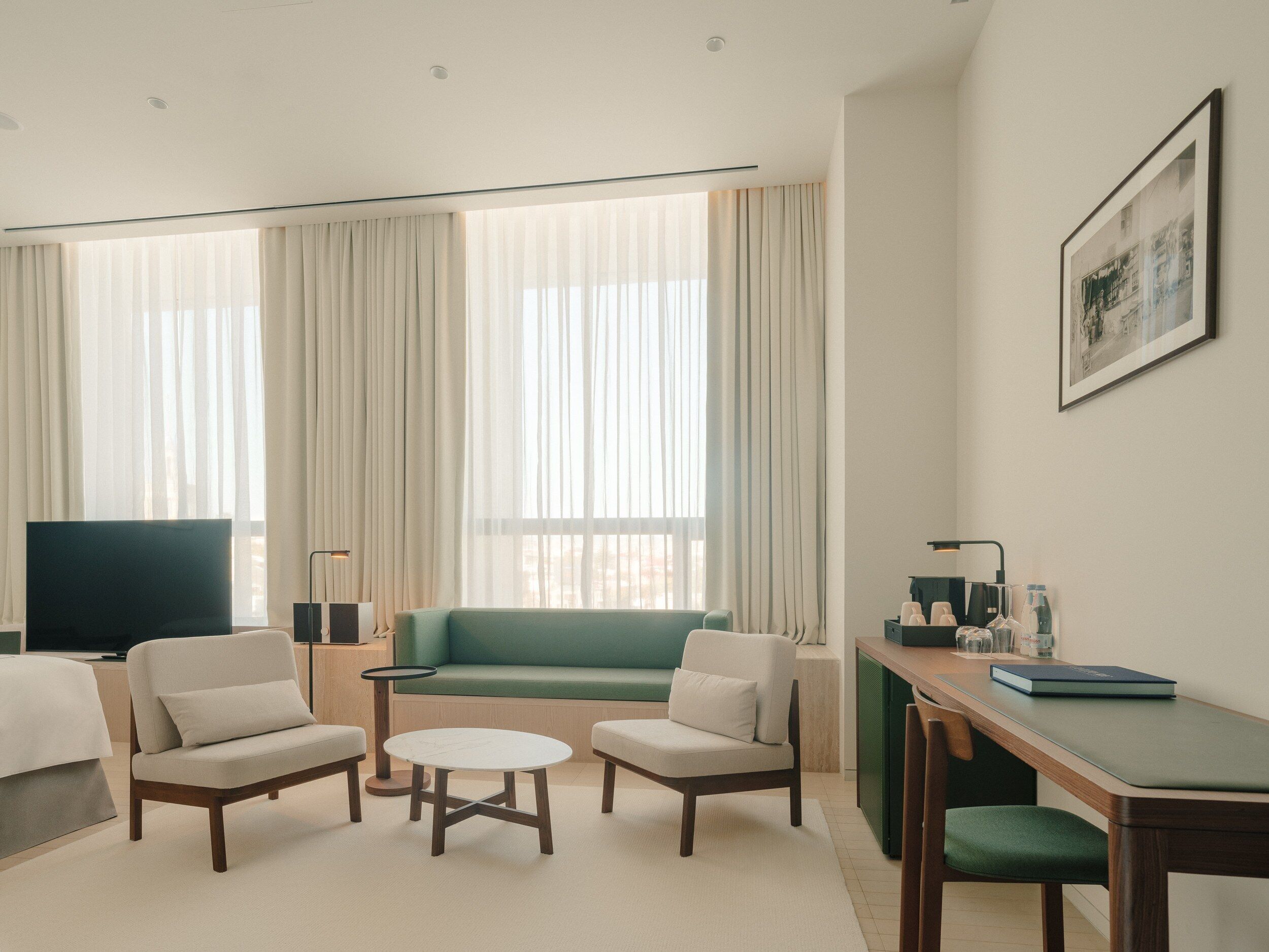 Une chambre d’hôtel moderne comprenant un canapé, deux fauteuils, une petite table ronde, un bureau avec une chaise, une télévision et un tableau encadré au mur.