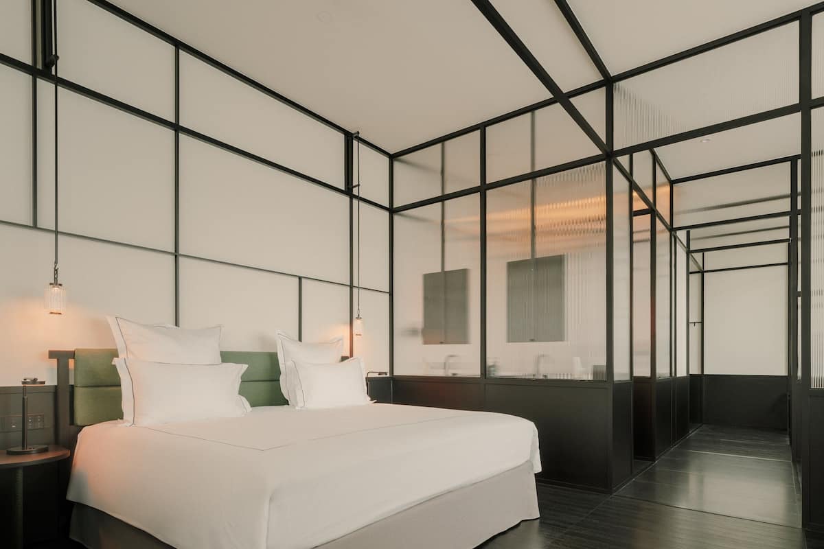 A modern hotel room with a large bed, bedside tables, and a view through a glass wall.