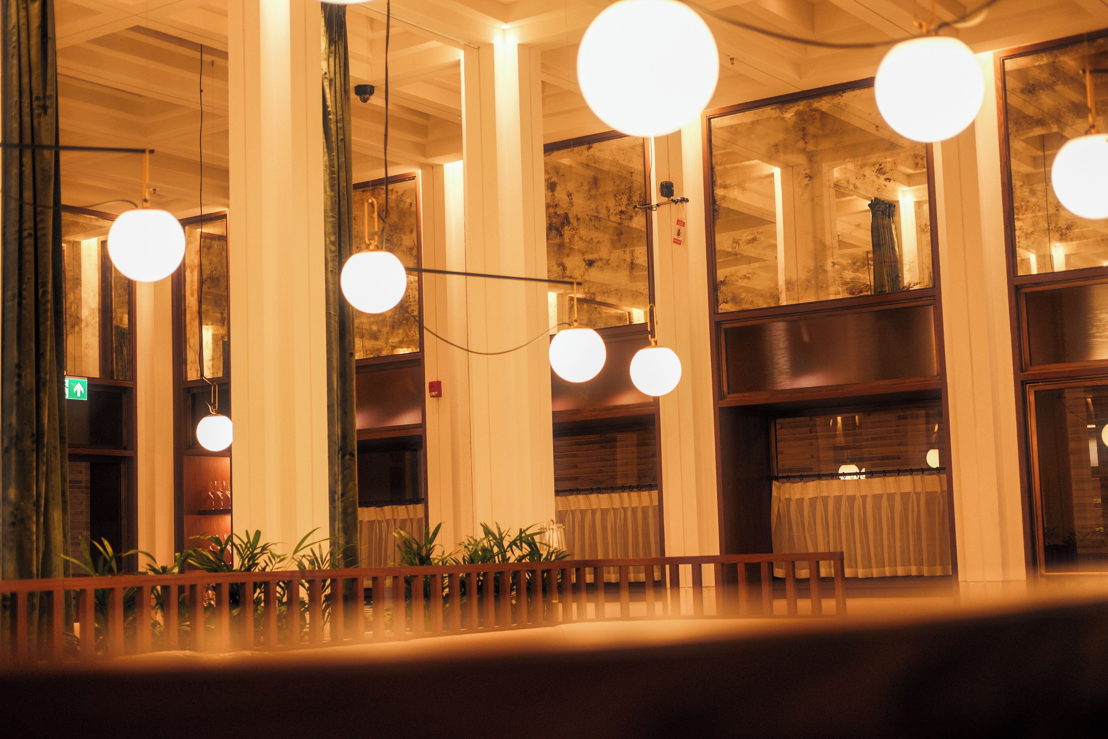 A hotel lobby with large columns, hanging spherical lights, and a wooden railing with plants.