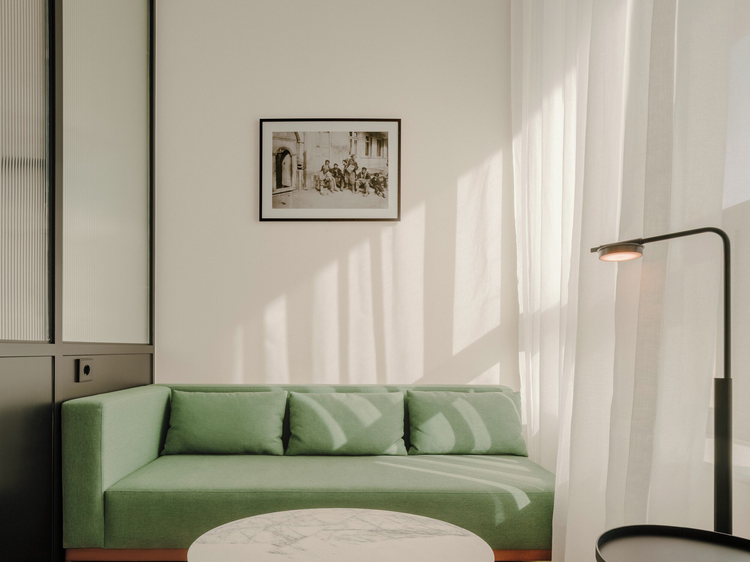 A green sofa with a framed picture on the wall, a floor lamp, and a window with blinds.