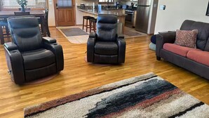 Un salón amplio con un sillón reclinable de cuero negro, un sofá con un cojín rojo y una alfombra a rayas de colores.