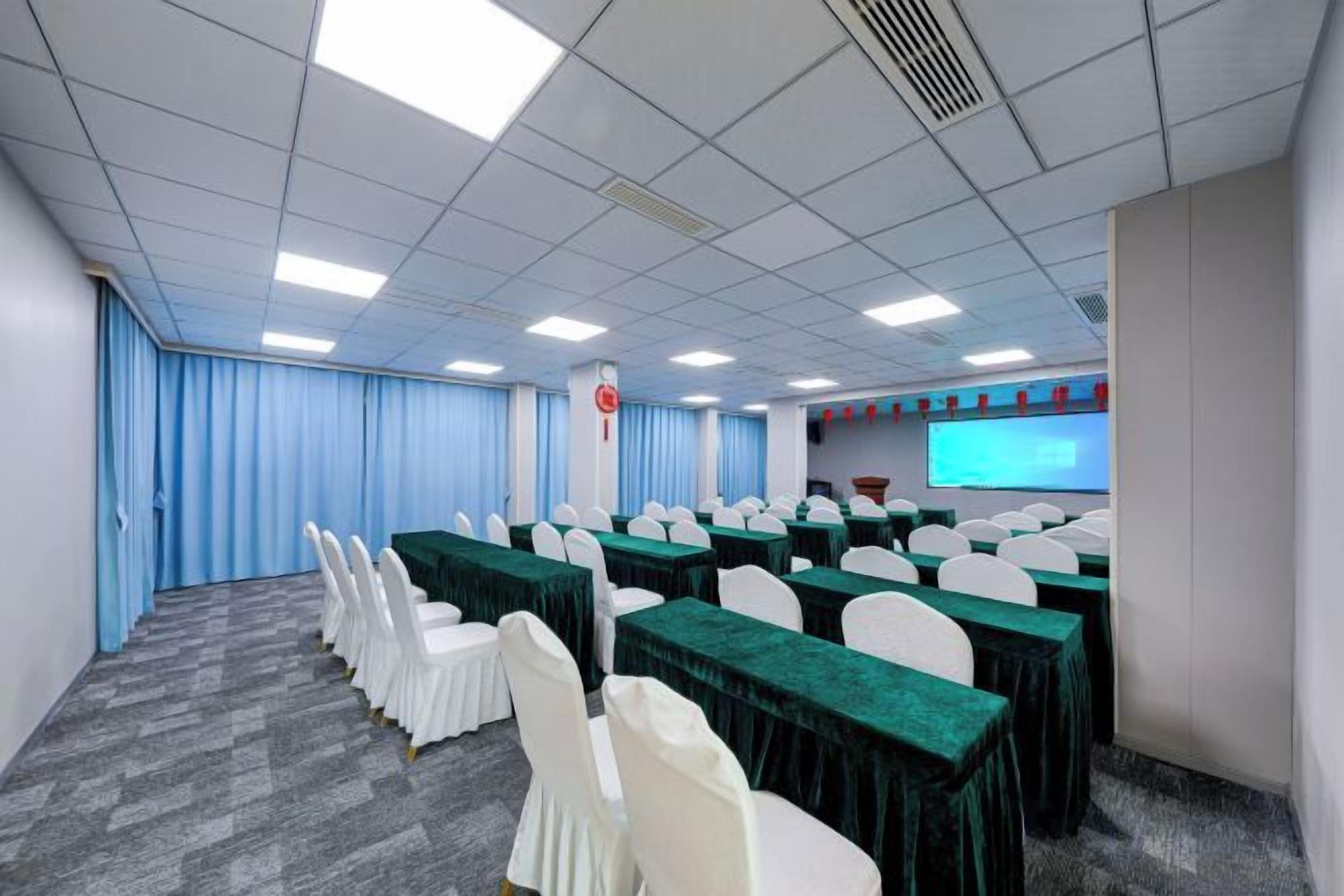 A conference room with rows of tables, chairs, a projector screen, and a blue curtain backdrop.
