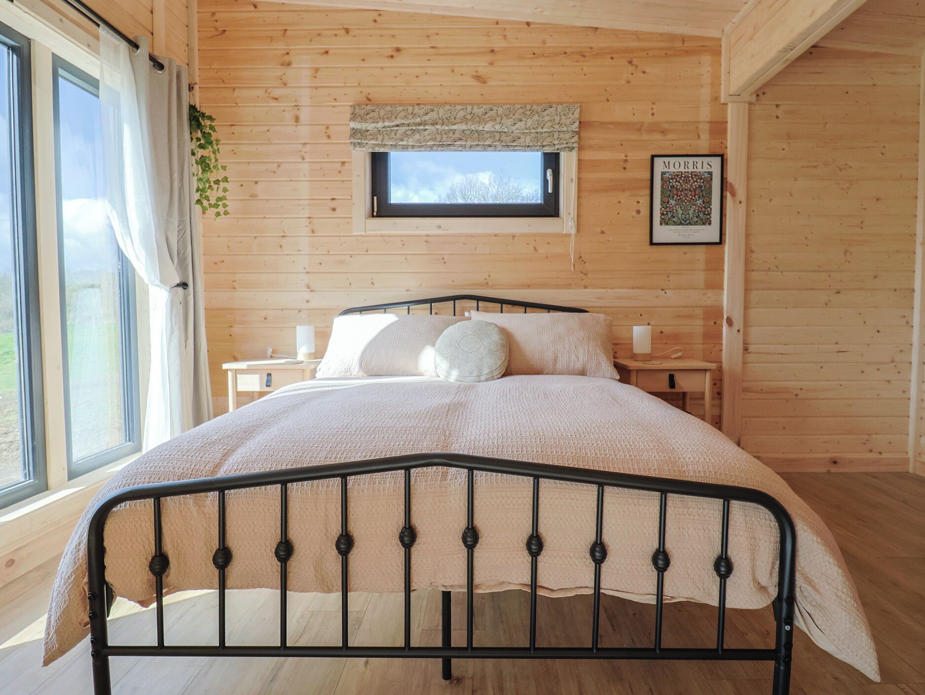 Un dormitorio de cabaña de madera con una cama, una ventana con cortinas, un ventanal con vistas y un cuadro en la pared.
