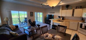 A living room with a sofa, coffee table, and dining table. There is a TV, kitchen area with cabinets, and a balcony with outdoor furniture.